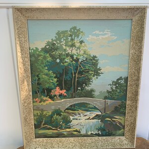 Puede incluir: Una pintura enmarcada de un paisaje con un puente de piedra sobre una cascada. La pintura est&aacute; hecha en un estilo de pintura por n&uacute;meros, con colores brillantes y un toque caprichoso.