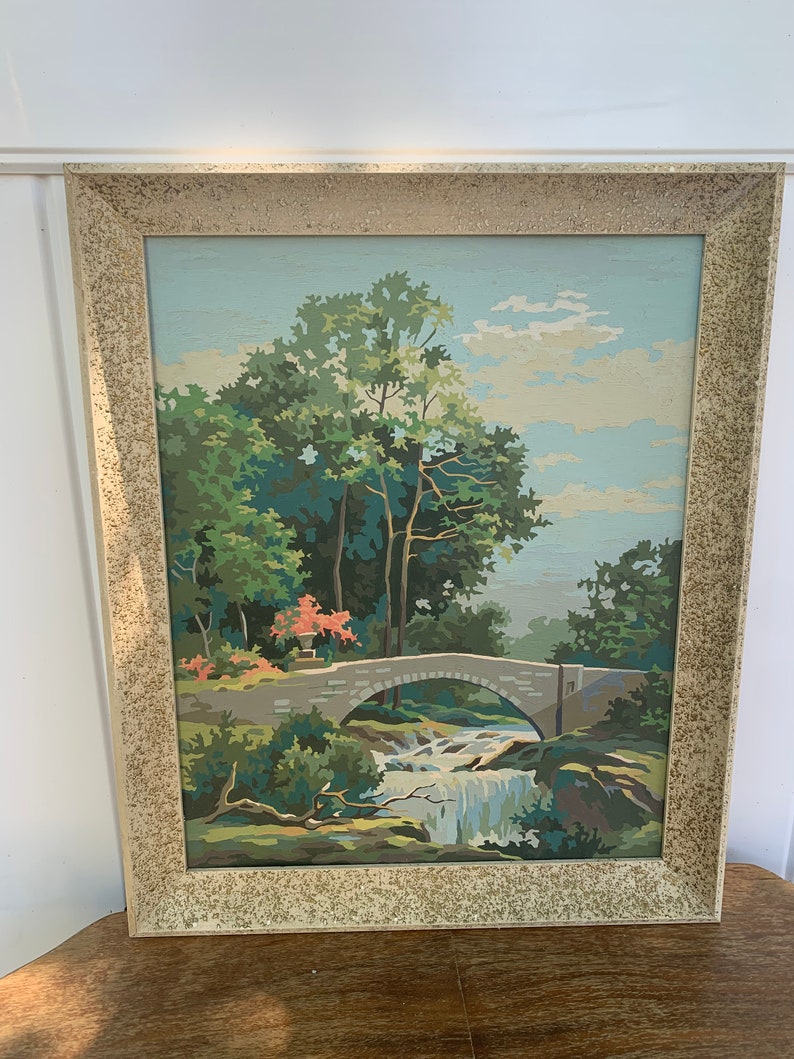 Puede incluir: Una pintura enmarcada de un paisaje con un puente de piedra sobre una cascada. La pintura est&aacute; hecha en un estilo de pintura por n&uacute;meros, con colores brillantes y un toque caprichoso.