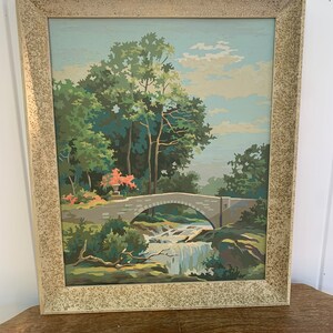 Puede incluir: Una pintura enmarcada de un paisaje con un puente de piedra sobre una cascada. La pintura est&aacute; hecha en un estilo de pintura por n&uacute;meros y presenta un cielo azul, &aacute;rboles verdes y un puente marr&oacute;n.
