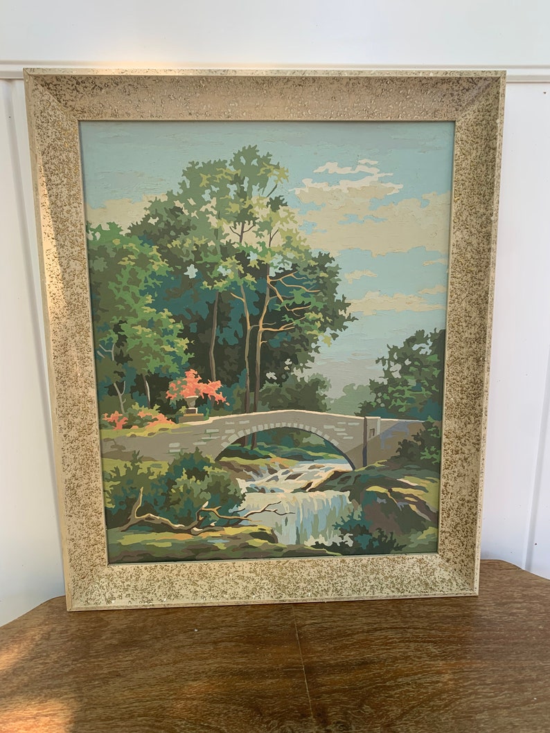 Puede incluir: Una pintura enmarcada de un paisaje con un puente de piedra sobre una cascada. La pintura est&aacute; hecha en un estilo de pintura por n&uacute;meros y presenta un cielo azul, &aacute;rboles verdes y un puente marr&oacute;n.