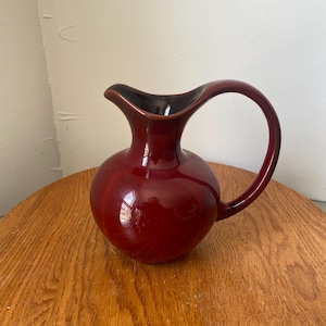 Peut inclure: Une cruche brillante de couleur marron foncé avec un intérieur sombre et une anse incurvée. La cruche est posée sur une surface en bois, mettant en valeur son corps arrondi et son design élégant. C'est un objet décoratif.