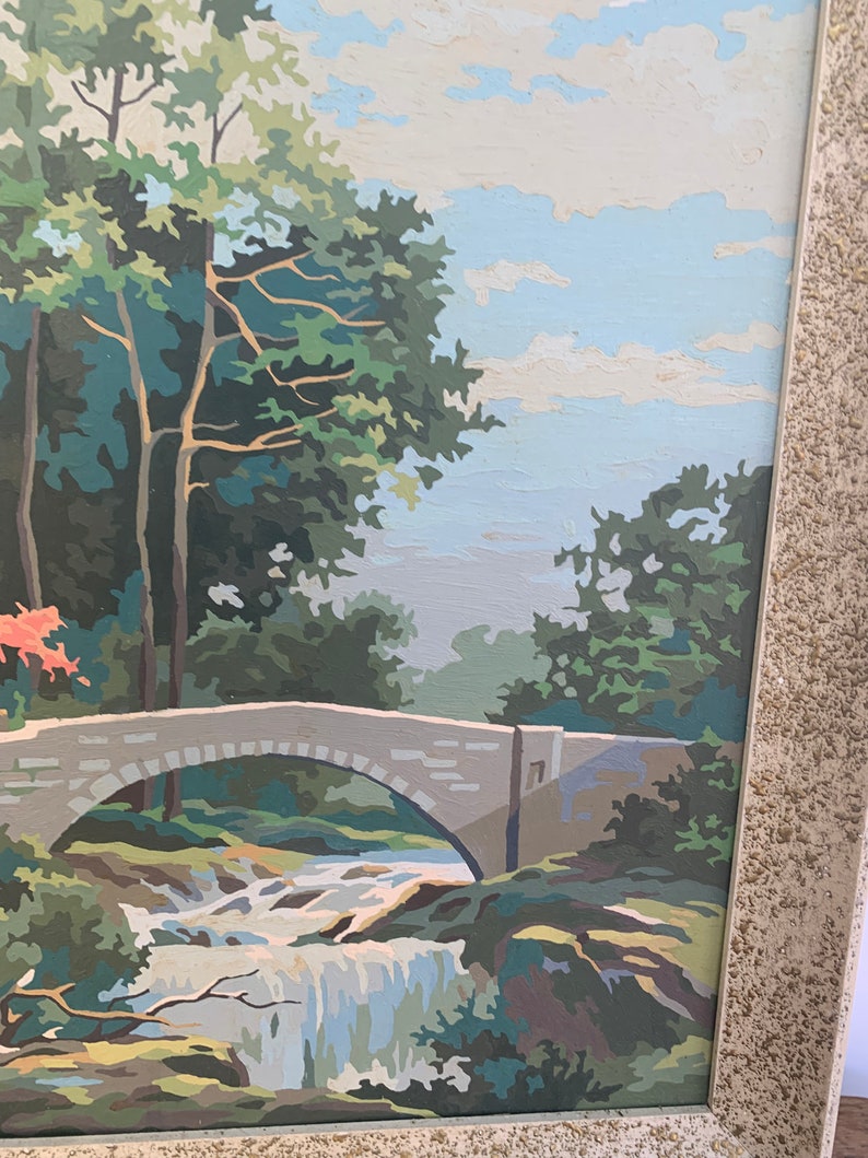 Puede incluir: Una pintura de un puente de piedra sobre una cascada que cae en un entorno boscoso. El cielo es azul con nubes blancas. Los &aacute;rboles son verdes y el agua es azul y blanca.