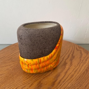 Könnte beinhalten: Eine kleine Keramikvase mit einzigartigem Design. Der obere Teil ist gesprenkelt dunkelbraun, während der untere Teil ein lebendiges, strukturiertes Muster aus Gelb, Orange und Rot aufweist. Der Innenraum ist weiß.