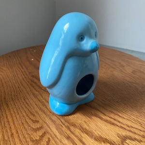 Puede incluir: Figura de pingüino de cerámica azul claro con una abertura redonda. El pingüino tiene un acabado liso y brillante, un pico azul pequeño y dos ojos pequeños. La figura está sobre una superficie de madera.