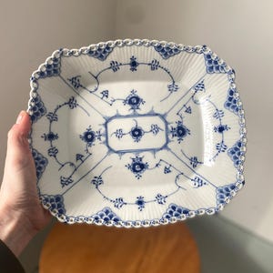 May include: A white porcelain serving dish with a blue floral design. The rectangular dish has a scalloped edge and a detailed blue pattern. The dish is held in a person's hand, showcasing its size and design.