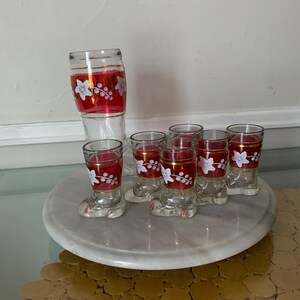 Puede incluir: Conjunto de seis vasos de chupito de vidrio transparente con una banda roja y dorada y un diseño floral blanco. Los vasos están dispuestos sobre una superficie de mármol blanco. Un vaso de vidrio transparente más grande con una banda roja y dorada y un diseño floral blanco se encuentra en el fondo.