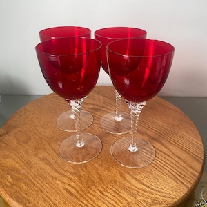 Puede incluir: Cuatro copas de vino tinto rubí con tallos transparentes y retorcidos y bases circulares. Las copas están dispuestas sobre una bandeja redonda de madera. Las copas tienen un diseño clásico y elegante.