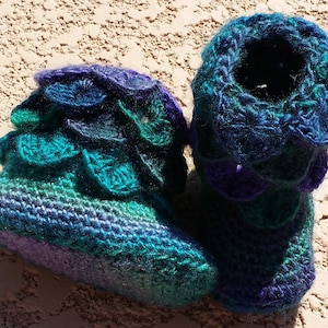May include: A pair of crocheted baby booties in shades of purple, teal, and blue. The booties have a scalloped edge and a textured design.