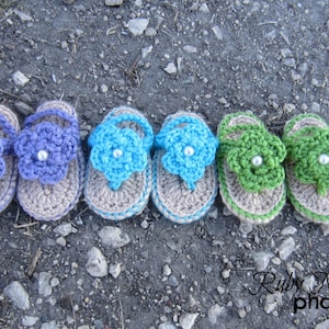 May include: Six pairs of crocheted baby sandals in various colors, including purple, blue, and green. Each sandal features a flower design with a pearl embellishment.