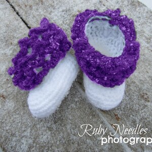 May include: Purple and white crocheted baby booties with a snowflake design. The booties are on a white surface with a wood grain texture.