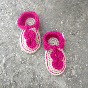 Puede incluir: Un par de sandalias de bebé de ganchillo en beige y rosa. Las sandalias tienen una correa que va alrededor del tobillo y una tira entre los dedos.