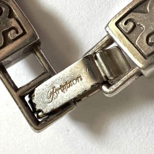 May include: Close-up of a silver-toned bracelet clasp. The clasp features an engraved design and the word "Brighton" is visible. The bracelet appears to be made of metal and has a decorative pattern.