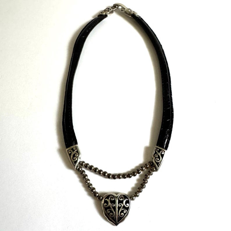 May include: A black leather necklace with a silver heart pendant. The pendant has an ornate design. The necklace has a silver clasp and silver accents. The necklace is approximately 16 inches long.