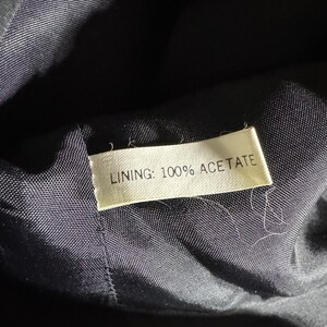 Pode incluir: Close-up de um interior de tecido escuro com uma etiqueta. A etiqueta diz "LINING: 100% ACETATE" em texto preto. O tecido parece ser de cor azul escuro ou preto.