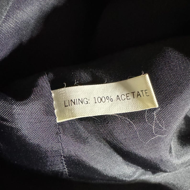 Pode incluir: Close-up de um interior de tecido escuro com uma etiqueta. A etiqueta diz "LINING: 100% ACETATE" em texto preto. O tecido parece ser de cor azul escuro ou preto.