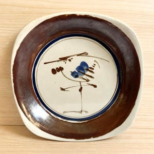 Pode incluir: Um prato quadrado de cerâmica com cantos arredondados, apresentando uma borda larga castanho-escura e um centro creme. O centro é decorado com uma borda circular fina azul-escura e um design abstrato pintado à mão com pinceladas castanhas e azuis.