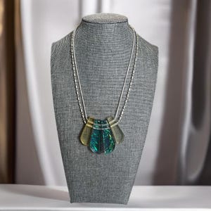 May include: A silver chain necklace with a multi-layered pendant made of green and gold iridescent glass. The pendant is shaped like a fan.