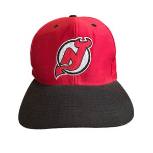 Può includere: Un cappello da baseball rosso e nero con il logo dei New Jersey Devils ricamato sul davanti.