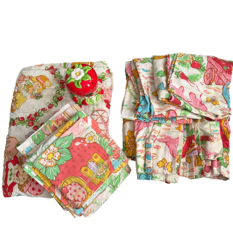 May include: A vintage Strawberry Shortcake bedding set with a white background and colorful floral and character designs. The set includes a quilt, pillowcases, and curtains.