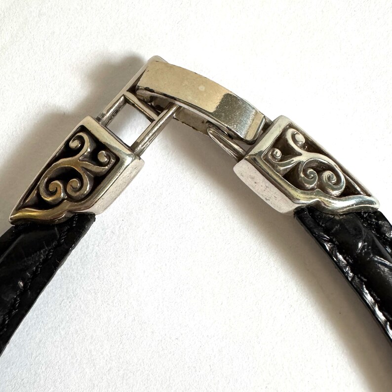 May include: Close-up of a silver bracelet clasp with ornate scrollwork on the ends. The bracelet band is black and appears to be made of leather. The clasp is rectangular and connects the two ends of the bracelet.