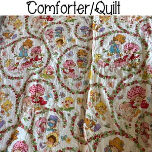 May include: A white comforter or quilt with a Strawberry Shortcake theme. The fabric features a repeating pattern of Strawberry Shortcake and her friends, including a red-haired girl with a strawberry on her head, a blonde girl with a blue dress, and a brunette girl with a pink dress. The characters are surrounded by strawberries, leaves, and flowers.