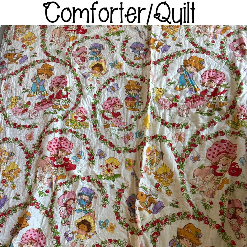 May include: A white comforter or quilt with a Strawberry Shortcake theme. The fabric features a repeating pattern of Strawberry Shortcake and her friends, including a red-haired girl with a strawberry on her head, a blonde girl with a blue dress, and a brunette girl with a pink dress. The characters are surrounded by strawberries, leaves, and flowers.