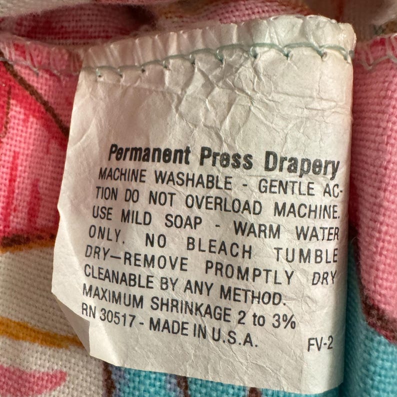 May include: A white care label with the text "Permanent Press Drapery" and instructions for washing and drying the fabric. The label includes the text "MACHINE WASHABLE - GENTLE ACTION DO NOT OVERLOAD MACHINE. USE MILD SOAP - WARM WATER ONLY. NO BLEACH TUMBLE DRY - REMOVE PROMPTLY DRY CLEANABLE BY ANY METHOD. MAXIMUM SHRINKAGE 2 to 3% RN 30517 - MADE IN U.S.A. FV-2"