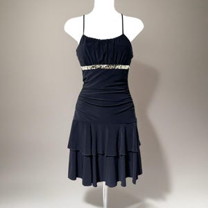 May include: A black dress with spaghetti straps and a fitted bodice. The dress features a jeweled band at the waist and a tiered, ruffled skirt. The dress is displayed on a white mannequin against a neutral background.