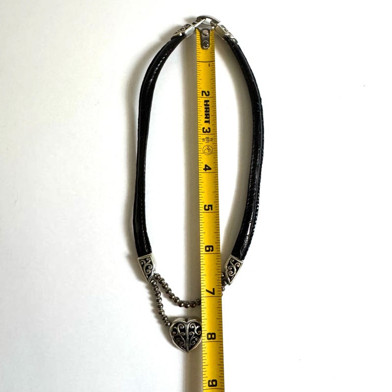 May include: Black leather necklace with silver-tone metal accents and a heart-shaped pendant. The necklace measures approximately 8 inches in length, as shown by the measuring tape.