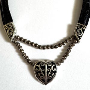 May include: A silver-toned necklace with a black heart-shaped pendant and a black leather strap. The pendant features an intricate, openwork design. The necklace has two strands of small, silver beads.