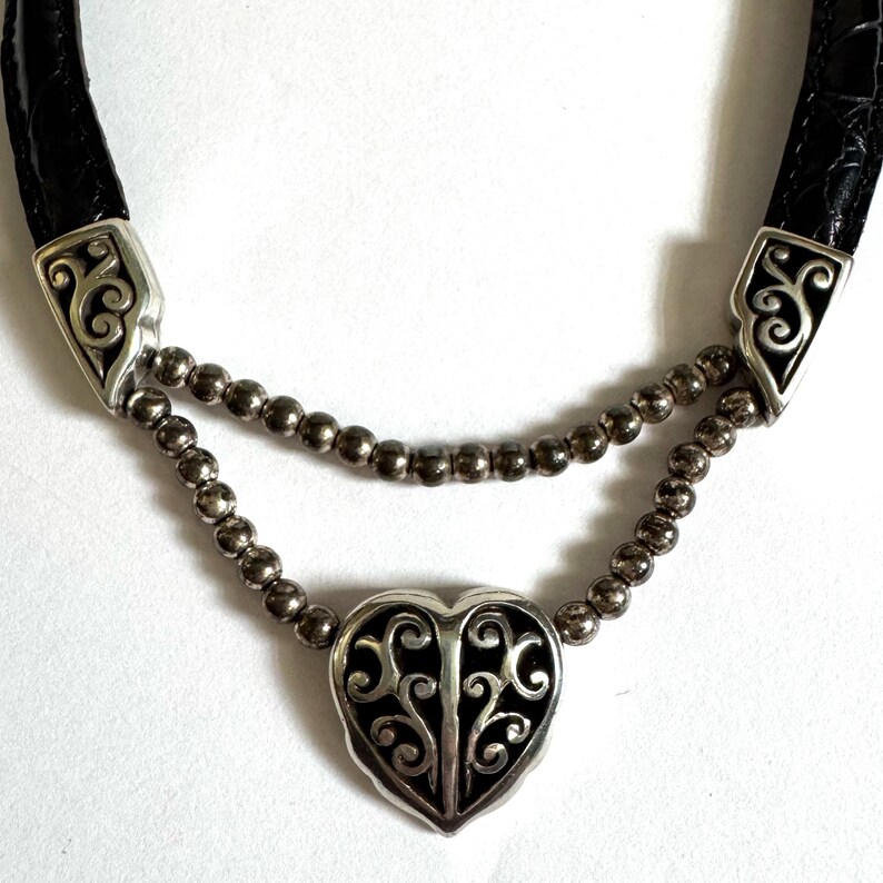 May include: A silver-toned necklace with a black heart-shaped pendant and a black leather strap. The pendant features an intricate, openwork design. The necklace has two strands of small, silver beads.