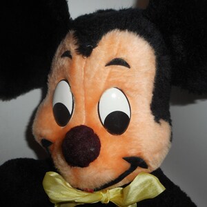 May include: Close-up of a vintage Mickey Mouse plush toy. The plush features a black head and ears, a peach-colored face, large white eyes with black pupils, a dark red nose, and a yellow bow tie. The toy is a classic Disney character.