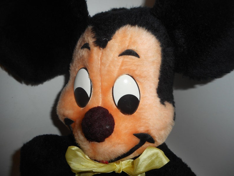 May include: Close-up of a vintage Mickey Mouse plush toy. The plush features a black head and ears, a peach-colored face, large white eyes with black pupils, a dark red nose, and a yellow bow tie. The toy is a classic Disney character.