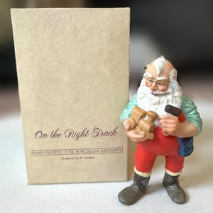 Puede incluir: Adorno de porcelana pintado a mano de Papá Noel sosteniendo un tren de madera y un martillo. Papá Noel viste un mono rojo, una camisa verde y botas grises. La caja dice "On the Right Track".