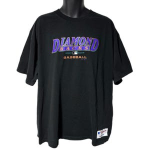以下が含まれることがあります： 紫とティール色の「Diamondbacks」の文字と、その下に「Authentic Collection Baseball」がプリントされた黒いTシャツ。半袖とクルーネックです。右下に小さな白いタグが見えます。