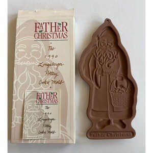 Puede incluir: Un cortador de galletas de terracota con forma de Papá Noel que lleva una cesta de regalos. El cortador está empaquetado en una caja con el texto "Father Christmas The 1990 Longaberger Pottery Cookie Mold".