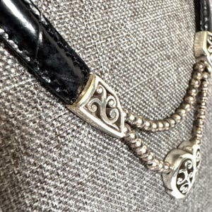 May include: Close-up of a black leather necklace with silver-toned metal accents. The necklace features decorative silver clasps with an ornate design. The background is a textured gray fabric.
