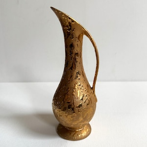 Könnte beinhalten: Goldfarbener, strukturierter Krug mit gebogenem Ausguss und Henkel. Die dekorative Vase hat einen bauchigen Boden und einen schlanken Hals. Die Oberfläche hat ein einzigartiges, unregelmäßiges Muster und wirkt vintage oder handgefertigt.