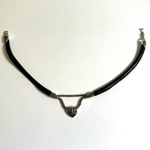 May include: Black choker necklace with a silver heart pendant. The necklace features a black leather cord with silver accents and a decorative heart-shaped pendant. The heart pendant has an intricate design. The necklace has a clasp closure.