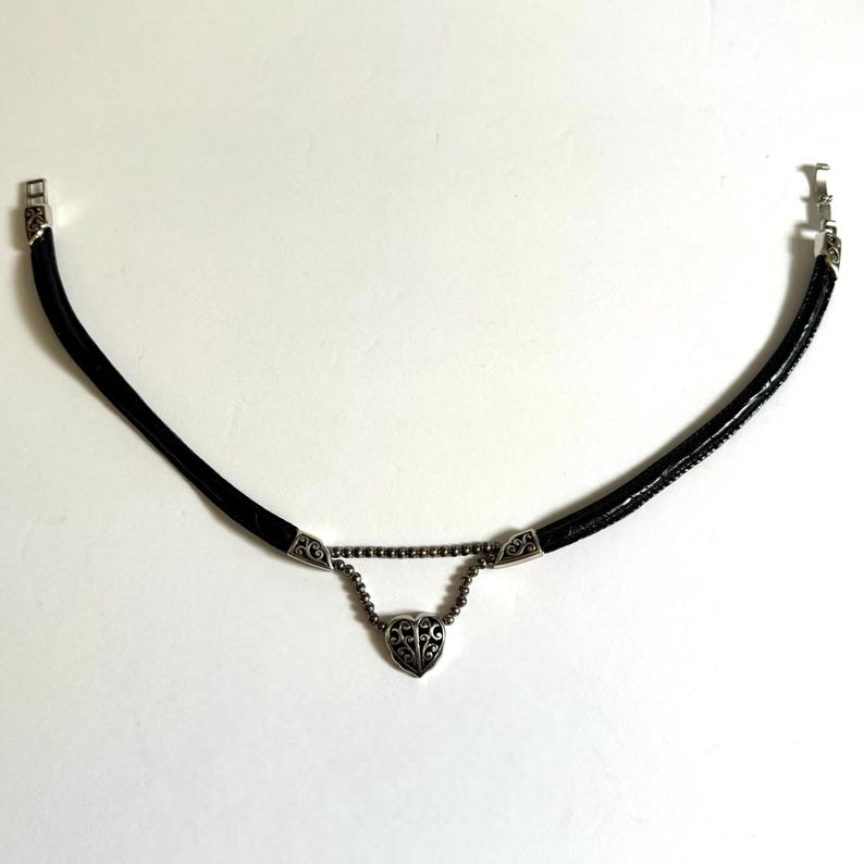 May include: Black choker necklace with a silver heart pendant. The necklace features a black leather cord with silver accents and a decorative heart-shaped pendant. The heart pendant has an intricate design. The necklace has a clasp closure.
