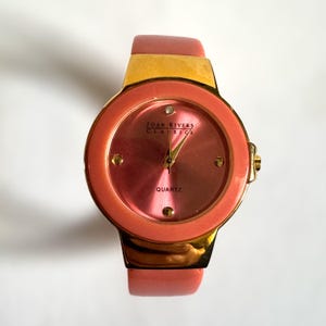 May include: A Joan Rivers Classics quartz watch with a pink face and a gold-tone bezel and band. The watch has a pink leather strap and a small diamond at the 12 o'clock position. The watch face has the word "QUARTZ" printed on it.