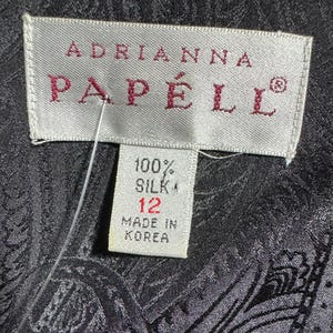Pode incluir: Close-up de uma etiqueta da Adrianna Papell. A etiqueta &eacute; branca com o nome da marca em bord&ocirc;. A etiqueta diz "100% Seda" e tamanho 12. A etiqueta tamb&eacute;m diz "Made in Korea". O fundo &eacute; um tecido escuro com estampa paisley.
