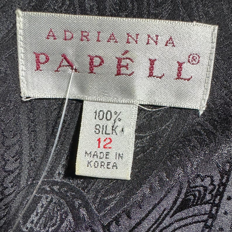 Pode incluir: Close-up de uma etiqueta da Adrianna Papell. A etiqueta &eacute; branca com o nome da marca em bord&ocirc;. A etiqueta diz "100% Seda" e tamanho 12. A etiqueta tamb&eacute;m diz "Made in Korea". O fundo &eacute; um tecido escuro com estampa paisley.