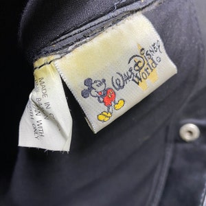 May include: A black fabric bag with a white label that says "Walt Disney World" and features a cartoon image of Mickey Mouse. The label is attached to the bag with white thread.