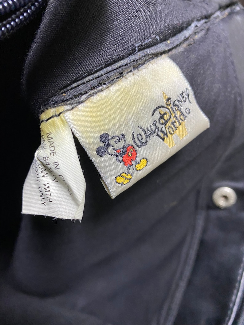 May include: A black fabric bag with a white label that says "Walt Disney World" and features a cartoon image of Mickey Mouse. The label is attached to the bag with white thread.
