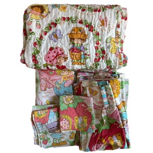 May include: A vintage Strawberry Shortcake bedding set featuring a quilted comforter and pillowcases. The comforter has a white background with a strawberry and leaf pattern and features images of Strawberry Shortcake and her friends. The pillowcases have a similar pattern with a pink and white color scheme.