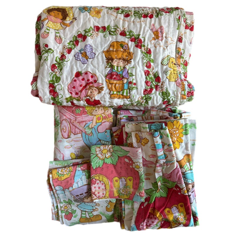 May include: A vintage Strawberry Shortcake bedding set featuring a quilted comforter and pillowcases. The comforter has a white background with a strawberry and leaf pattern and features images of Strawberry Shortcake and her friends. The pillowcases have a similar pattern with a pink and white color scheme.