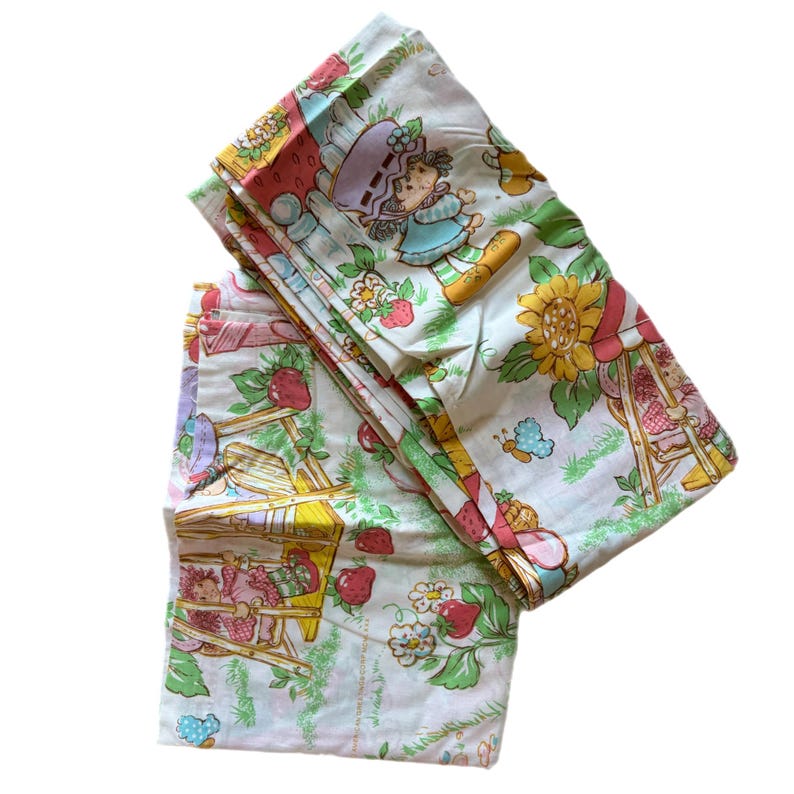 May include: A set of two vintage Strawberry Shortcake twin bed sheets featuring a white background with a green, yellow, and pink floral pattern. The sheets feature Strawberry Shortcake and her friends.