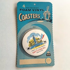 May include: A package of four mar-resistant foam vinyl coasters. The coasters are round with a white top featuring a Gettysburg, Pennsylvania, memorial design. The package is light blue with the word "COASTERS" in large, stylized letters.