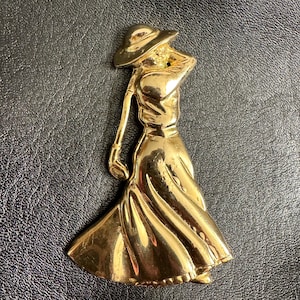 Puede incluir: Broche de metal dorado con la forma de una mujer con sombrero y vestido largo, sosteniendo un palo de golf. El broche tiene un acabado pulido y probablemente sea vintage.